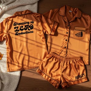May include: Orange satin pajama set with black trim. The top has a pocket with a megaphone graphic and the word "Cheer". The back of the top says "Dreaming of ZERO Deductions". The shorts say "Henley".