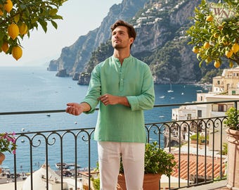 May include: A light green linen shirt with a mandarin collar and long sleeves, worn with white trousers. The man is on a balcony overlooking the sea, with mountains and buildings in the background. Lemon trees are visible.