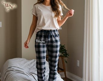May include: Black and gray plaid pajama pants with the word "Princess" in white script. The pants have a drawstring waist. The person is wearing a light beige t-shirt.