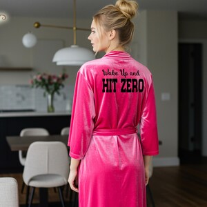 May include: A bright pink robe featuring the text "Wake Up and HIT ZERO" on the back. The robe is made from a soft, velvety fabric and has a tie closure. The image focuses on the back of the robe, showcasing the prominent text.