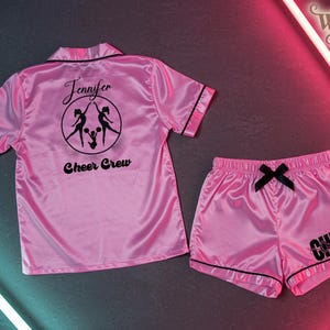 May include: A pink satin pajama set with black trim. The top has a collar and short sleeves, with "Jennifer" and "Cheer Crew" printed on the back. The shorts have a black bow and "Cheer Coach" printed on the leg.