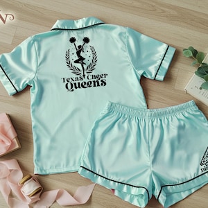 May include: A light blue satin pajama set with black trim. The top features the text "Texas Cheer Queens" and a cheerleader graphic. The shorts have a megaphone graphic and the name "Dianna".