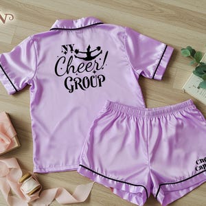 May include: Lilac satin pyjama set with black trim. The top has short sleeves and the text "NY Cheer! GROUP" on the back. The shorts have "Cheer Captain" printed on the leg. The set is displayed on a light wood surface.