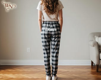 May include: Black and gray plaid pajama pants with the name "LAUREN" printed in white. The pants have an elastic waistband and are cuffed at the ankles. The person is wearing a beige t-shirt and white sneakers.