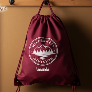 May include: A burgundy drawstring backpack with a white circular logo featuring a mountain and lake scene. The text "Wilderness Division" and the name "Amanda" are printed on the bag. The bag is hanging on a wooden rack.