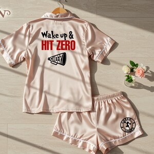 May include: A pale pink satin pajama set with white edging. The top features the text "Wake up & Hit Zero" and a megaphone graphic. The shorts have a cheerleader logo. The set is hanging on a wooden hanger.