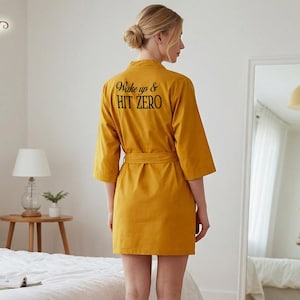 Peut inclure: Peignoir jaune moutarde avec une ceinture assortie. Le dos du peignoir porte l'inscription "Wake up & Hit Zero" en noir. Le peignoir a des manches larges et arrive au niveau des genoux.