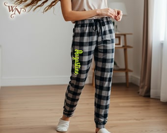 May include: Black and gray plaid pajama pants with a drawstring waist. The name "Augustine" is printed vertically in a bright yellow script on the left leg. The pants are made of a soft material and are cuffed at the ankles.