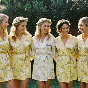 May include: Five women wearing floral print robes with matching flower crowns. The robes are yellow with white floral patterns. One robe is white with a floral pattern and the word "Bride" embroidered on it. The women are smiling and standing in front of green foliage.
