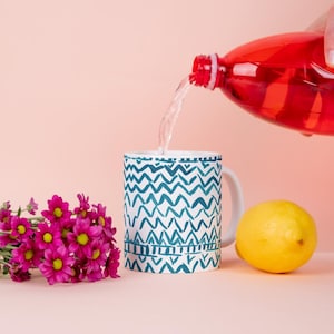 May include: A white ceramic mug with a teal zigzag pattern is being filled with liquid from a red bottle. A yellow lemon and a bouquet of pink flowers with yellow centers are next to the mug. The background is a solid pink.