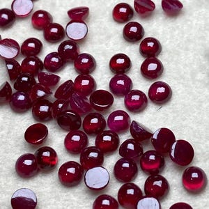 Könnte beinhalten: Eine Ansammlung kleiner, runder, polierter Rubin-Edelsteine. Die Edelsteine haben eine tiefrote Farbe, wobei einige eine flache, unpolierte Rückseite aufweisen. Die Edelsteine sind auf einer weißen Oberfläche verstreut, einige sind halbiert.