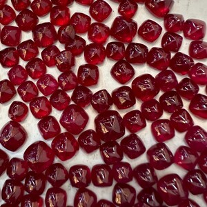 May include: A scattering of square-cut, deep red gemstones. The gems are faceted and reflect light, creating a sparkling effect. They are arranged on a light surface, highlighting their rich colour and polished appearance. A luxurious collection.