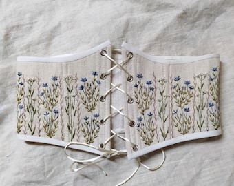 May include: Cream-colored corset with embroidered blue and green floral design. The corset features a white trim and a lace-up front with silver eyelets. The floral embroidery is detailed and covers the top and bottom edges of the corset.