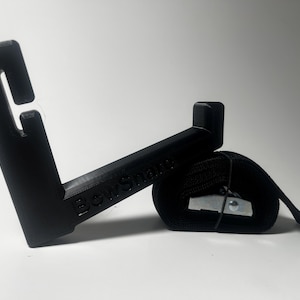 Puede incluir: Dispositivo BowSnake negro con gancho y correa. El dispositivo tiene el texto "BowSnake" en el lateral. La correa es negra y tiene una hebilla metálica. El dispositivo está diseñado para sujetar algo.