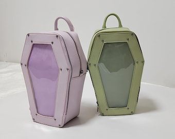 May include: Two coffin-shaped backpacks. One is light pink with a purple window, the other is light green with a clear window. Both have a top handle, zipper closure, and metal accents. The bags are on a white surface.