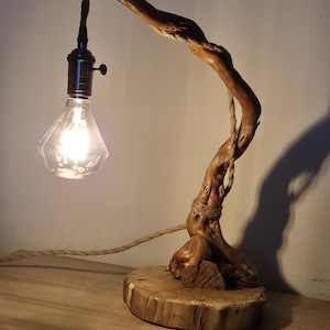 Pode incluir: Um candeeiro de mesa com uma peça natural e curva de madeira flutuante como suporte principal, montado numa base circular de madeira. Uma corda grossa de cor bege envolve a madeira, suspendendo um casquilho de metal preto com uma lâmpada.