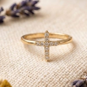 May include: A gold-colored ring featuring a cross design encrusted with small, sparkling stones. The ring is set against a neutral, textured background, with hints of lavender and dried flowers in the periphery. The cross is the focal point.