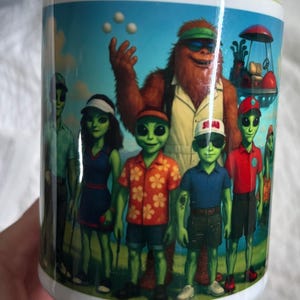 May include: White ceramic mug featuring a whimsical illustration of a golf-themed scene with green-skinned aliens and a Sasquatch. The aliens are dressed in golf attire, and the mug has a green rim. The word "SQUAD" is on a hat.