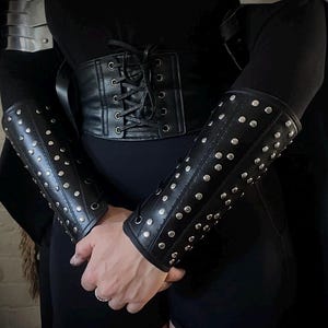 May include: Black leather corset and studded arm guards. The corset has a lace-up front. The arm guards are adorned with silver studs. The overall aesthetic suggests a medieval or fantasy theme.