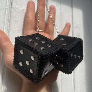 May include: Two black leather dice connected by a silver-toned metal chain. Each cube features silver metal studs arranged to represent the pips on a die, with black stitched edges along the seams of the leather construction.