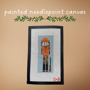 May include: Painted needlepoint canvas with a cross-stitch design of a person in hunting attire, including an orange vest and hat, and camouflage trousers. The canvas is framed in black and includes the initials "S+B".