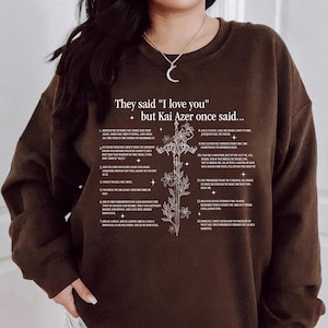 Może przedstawiać: Ciemnobrązowa bluza z napisem "They said 'I love you' but Kai Azer once said..." w kolorze białym. Na przodzie znajduje się kwiatowy wzór krzyża. Bluza to swobodny top z długim rękawem.