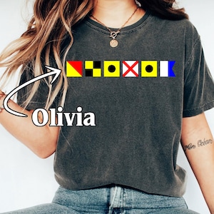 May include: Dark gray t-shirt featuring a row of colorful nautical signal flags. The name "Olivia" is printed in white below an arrow pointing to the flags. The word "PEPPER" is printed in white at the bottom of the image.