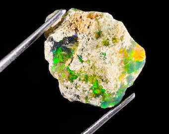 May include: A raw, unpolished opal gemstone featuring a rough, textured surface with visible inclusions. The stone displays vibrant flashes of green, yellow, and orange play-of-color against a translucent and earthy, beige-toned matrix base.