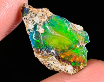 May include: A raw, unpolished opal gemstone featuring a vibrant play of color with intense flashes of neon green, electric blue, and warm orange. The stone has an irregular, jagged shape with visible rough, earthy matrix rock along its edges.