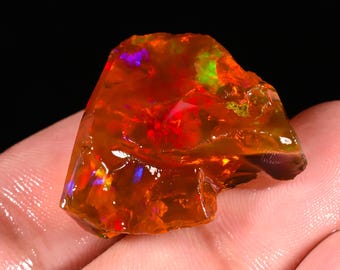 May include: A rough, uncut opal gemstone with a fiery orange and red base color. The stone displays flashes of green, blue, and purple iridescence. The opal is held in a hand against a black background.