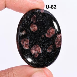 May include: An oval-shaped polished gemstone featuring a deep black base color with scattered, irregular dark red and brownish inclusions throughout the surface. The stone is marked with the text U-82 in the upper right corner of the image.