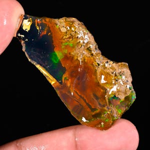 May include: A rough, uncut opal gemstone with a vibrant display of colors. The opal features a mix of orange, green, and black hues, with a translucent quality. The stone is held between fingers, showcasing its natural, raw form.
