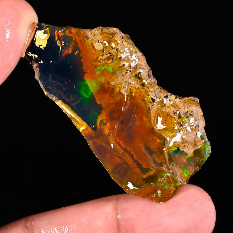 May include: A rough, uncut opal gemstone with a vibrant display of colors. The opal features a mix of orange, green, and black hues, with a translucent quality. The stone is held between fingers, showcasing its natural, raw form.