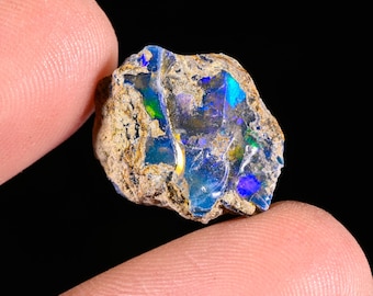May include: A raw, unpolished opal gemstone featuring a rough, earthy brown exterior texture and a vibrant, iridescent interior display of electric blue, green, and purple flashes of color set against a dark, natural stone matrix background.