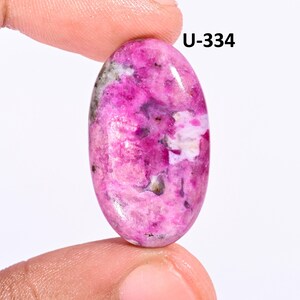Puede incluir: Una piedra preciosa cabujón de forma ovalada que presenta una paleta de colores magenta y rosa vibrantes con inclusiones moteadas blancas y gris oscuro en toda la superficie. La piedra está marcada con el texto U-334 en la esquina superior.