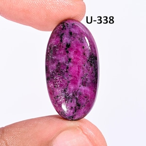 May include: An oval-shaped cabochon gemstone featuring a vibrant magenta and deep pink base color with scattered black mineral inclusions throughout the surface. The stone is marked with the text U-338 in the upper right corner of the frame.
