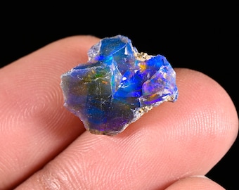 May include: A raw, uncut opal gemstone displaying vibrant flashes of electric blue, deep purple, and subtle green iridescence across its multifaceted, translucent surface. The stone features jagged, natural edges and a polished, reflective texture.