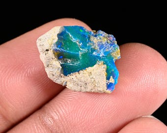 May include: A raw, unpolished opal gemstone featuring a vibrant, iridescent blue and green center surrounded by a rough, beige-colored rocky matrix. The stone displays a natural, irregular shape with a glossy, reflective surface texture.