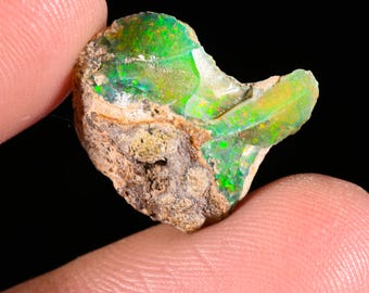 May include: A raw, unpolished opal gemstone featuring a vibrant play of color with bright green and yellow flashes. The stone has a rough, textured, earthy brown matrix on one side, contrasting with the smooth, translucent, iridescent surface.