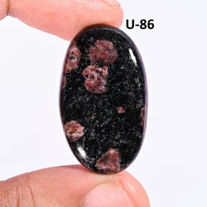 May include: An oval-shaped polished gemstone featuring a deep black base color with scattered, irregular dark red inclusions throughout the surface. The stone is marked with the text U-86 in the upper right corner, displaying a smooth finish.
