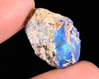 May include: A raw, unpolished opal gemstone featuring a rough, tan-colored exterior matrix and a vibrant, iridescent blue and green play-of-color visible within the fractured, crystalline interior sections of the stone.