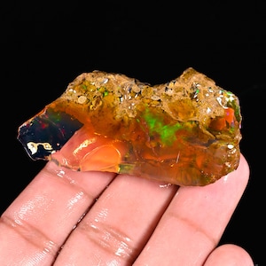 May include: A rough, uncut opal gemstone with vibrant colors. The opal displays a mix of orange, green, and red hues, with a rough, textured surface. The gemstone is held in a hand against a black background.