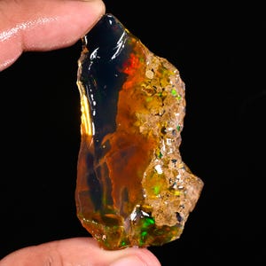 May include: A rough, uncut opal gemstone with a dark, translucent body. The stone displays flashes of red, green, and yellow, with a rough, textured surface on one side. The opal is held between fingers against a black background.