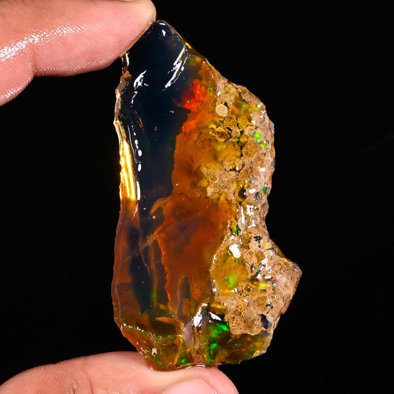 May include: A rough, uncut opal gemstone with a dark, translucent body. The stone displays flashes of red, green, and yellow, with a rough, textured surface on one side. The opal is held between fingers against a black background.
