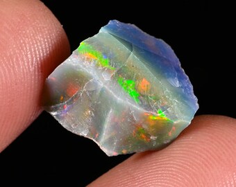 May include: A raw, uncut opal gemstone featuring a translucent blue base with vibrant flashes of green, orange, and red play-of-color. The stone has an irregular, jagged shape with visible natural fractures and a polished, reflective surface texture.