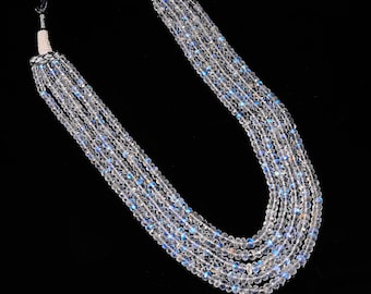 May include: A multi-strand necklace featuring five rows of faceted, translucent moonstone beads. Each bead displays a subtle iridescent blue sheen. The strands are secured at the top with white seed bead accents and a black cord closure.
