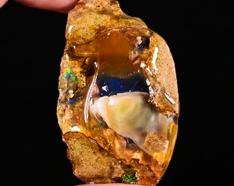 May include: A rough, uncut opal gemstone with a mix of colors. The opal features a base of tan and brown with areas of blue, green, and yellow. The stone is held between fingers against a black background.