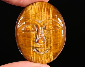 May include: An oval-shaped tiger's eye gemstone carved with a serene face featuring closed eyes, a defined nose, and a subtle smile. The stone displays a polished, glossy finish with characteristic golden-brown vertical chatoyant bands.