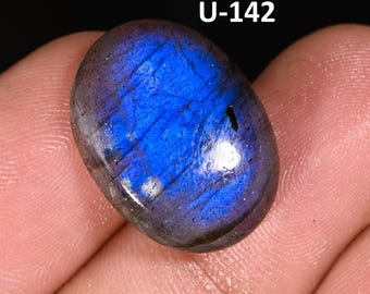 May include: An oval-shaped labradorite gemstone featuring a polished surface with a vibrant blue and purple iridescent flash. The stone displays natural internal fractures and inclusions, with the text U-142 printed in white above the stone.