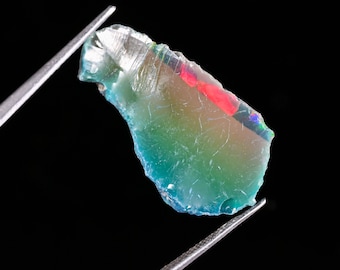 May include: A raw, translucent opal gemstone with a jagged, irregular shape held by metal tweezers. The stone displays a vibrant play of color, featuring shifting hues of teal, green, and blue, with a distinct, bright red flash along the top edge.
